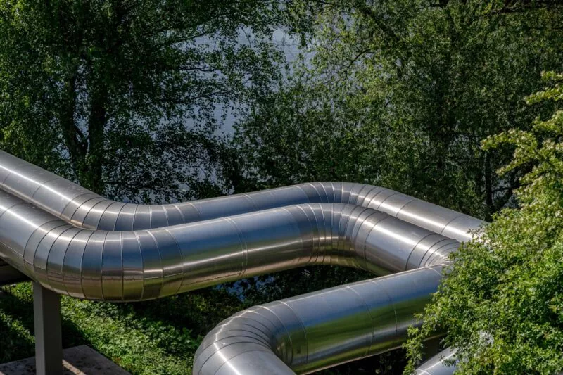 Ein großes, geschwungenes Rohr für die Fernwärme im Sonnenlicht vor einem grünen Baum.